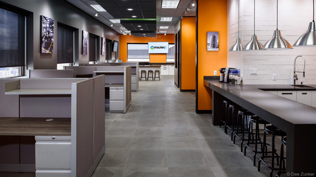 Modern industrial office interior with open workstations, orange accent wall, kitchen bar seating, and presentation screen in Houston corporate renovation