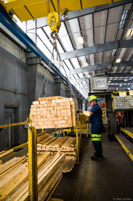 Worker in hard hat and hi-vis gear guides overhead crane lifting stacked lumber inside Houston industrial warehouse facility.