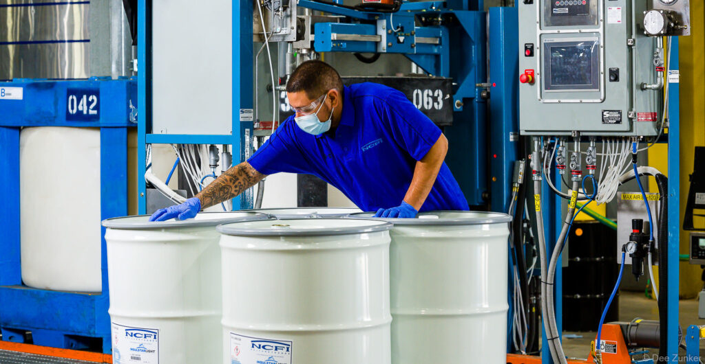 Worker in blue uniform, mask, and gloves positions white chemical drums at industrial manufacturing facility, Houston commercial photography.