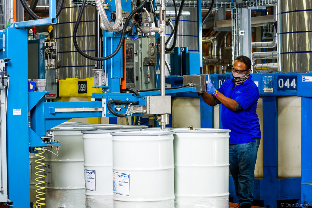 Worker in PPE mask and gloves operates drum-filling machinery at an industrial chemical manufacturing facility in Houston.