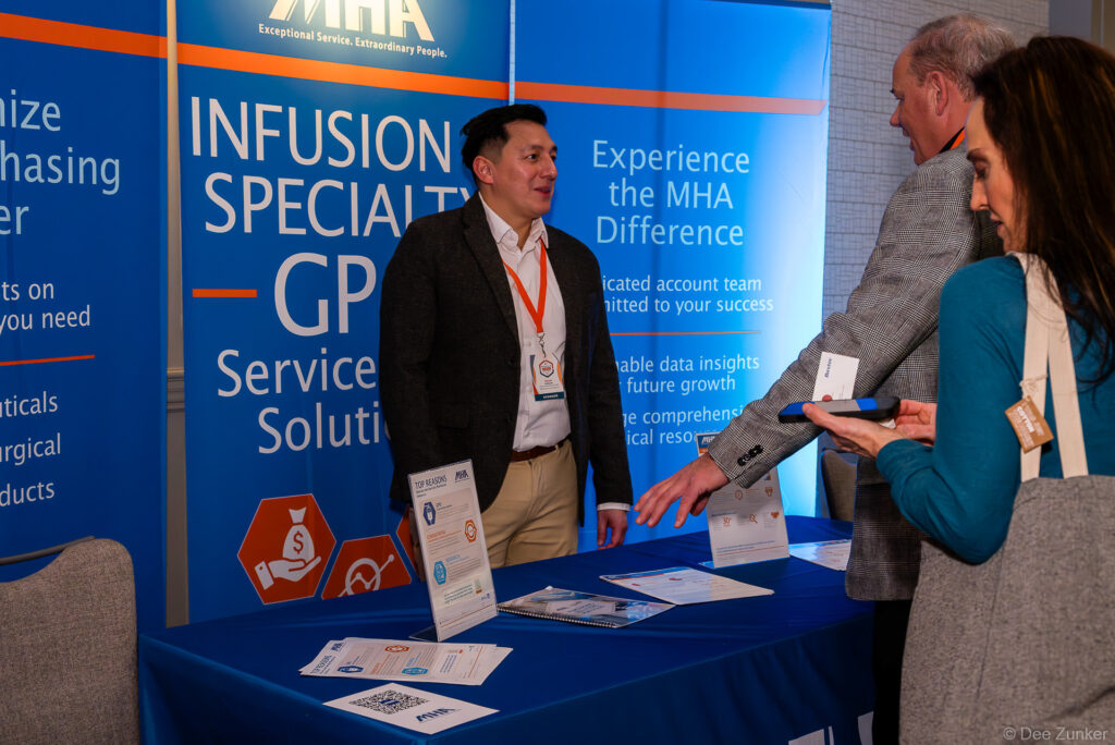 Conference exhibitor booth rep speaks with two attendees at an infusion specialty GPO services display table with branded banners.