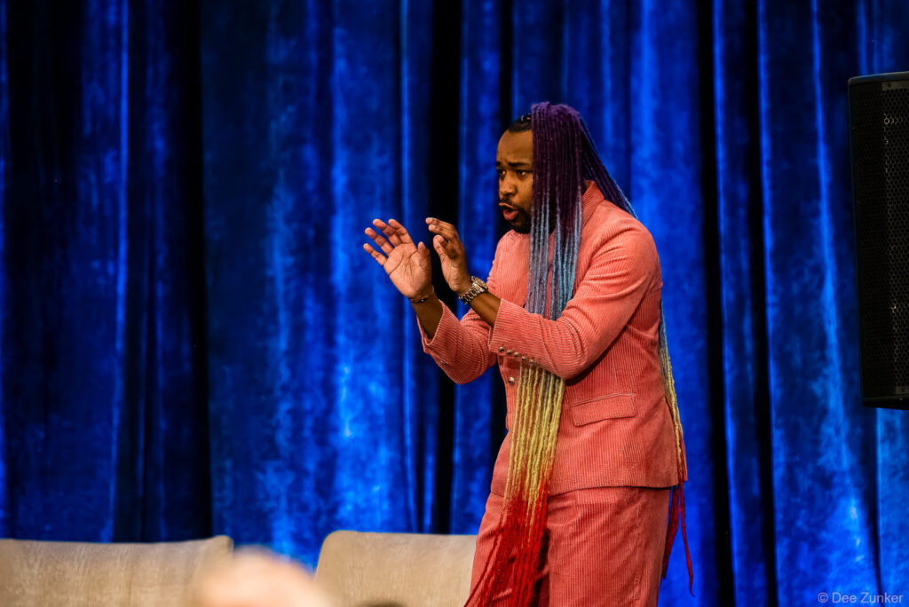 Keynote speaker in pink corduroy suit with colorful braids gestures on stage at INTA Leadership Meeting conference event