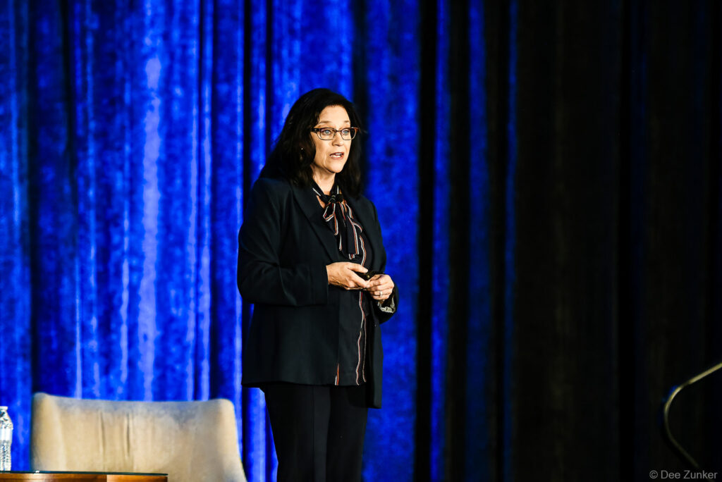 Female keynote speaker addresses INTA Leadership Meeting audience on stage before blue drape backdrop, holding presentation remote.