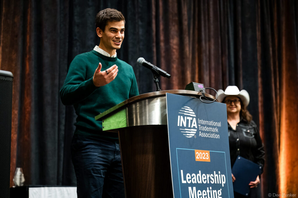 Young male speaker at INTA Leadership Meeting podium gestures while addressing audience, woman in cowboy hat stands behind him on stage.