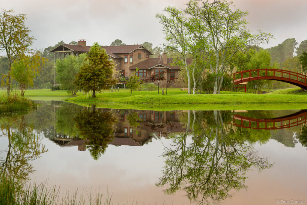 Luxury Houston estate exterior with red arched bridge, manicured grounds, and still pond reflection on overcast day.