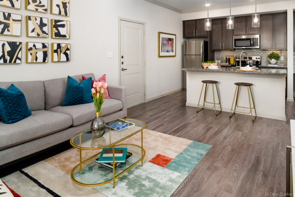 Open-concept apartment living room and kitchen with gray sofa, teal pillows, gold coffee table, and dark wood cabinetry