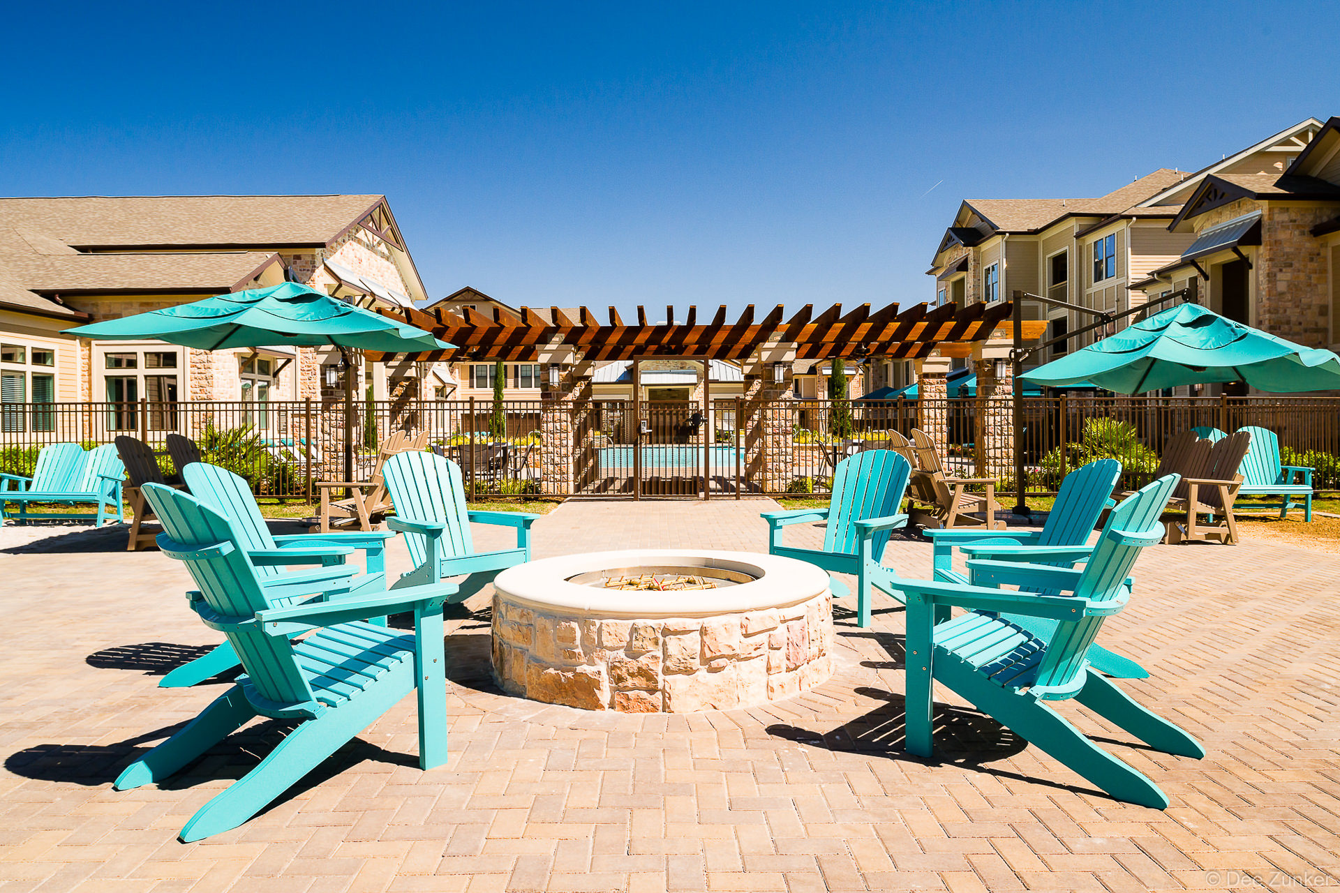 Turquoise Adirondack chairs surrounding a stone fire pit patio at a Katy Texas apartment complex with pool pergola beyond