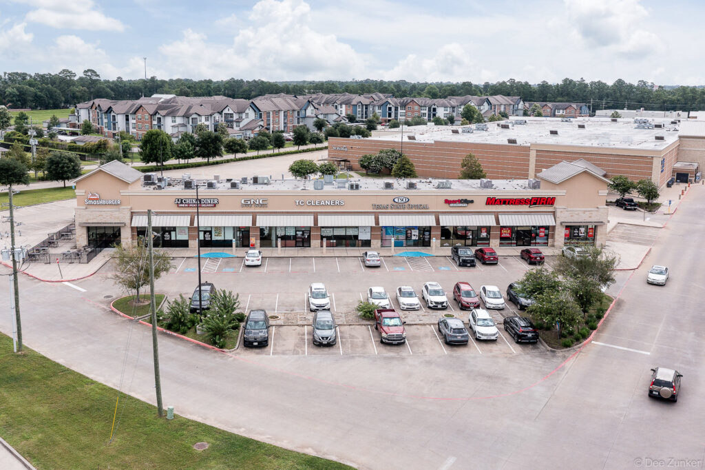 Aerial drone view of a retail strip center in Kingwood TX featuring storefronts, parking lot, and surrounding residential apartments.