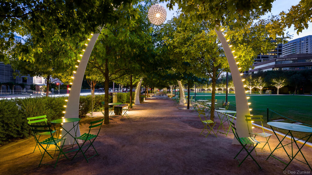 Illuminated arched sculptures and green bistro chairs line a tree-lined pathway in Klyde Warren Park, Houston, at dusk