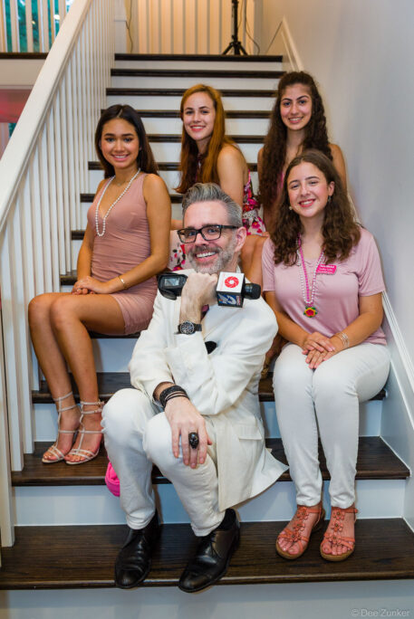 Man in white suit holds TV microphone beside four young women on staircase at Houston corporate event
