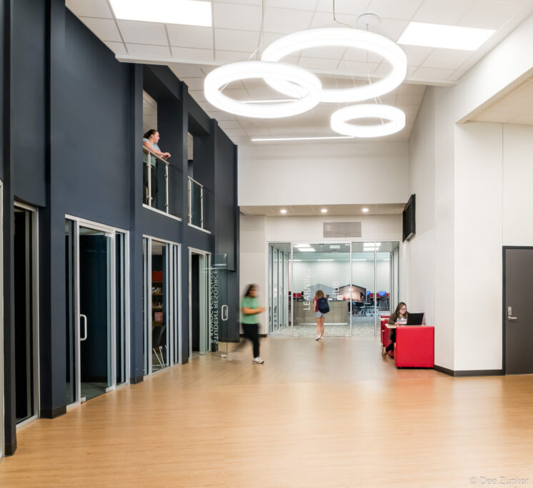 Two-level college student center interior hall with circular pendant lights, wood floors, red seating, and a glass-walled learning hub in Baytown TX