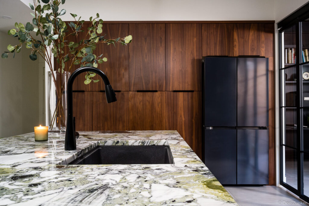 Luxury kitchen interior with green marble island, matte black faucet, walnut cabinetry, and black French-door refrigerator in Houston home.