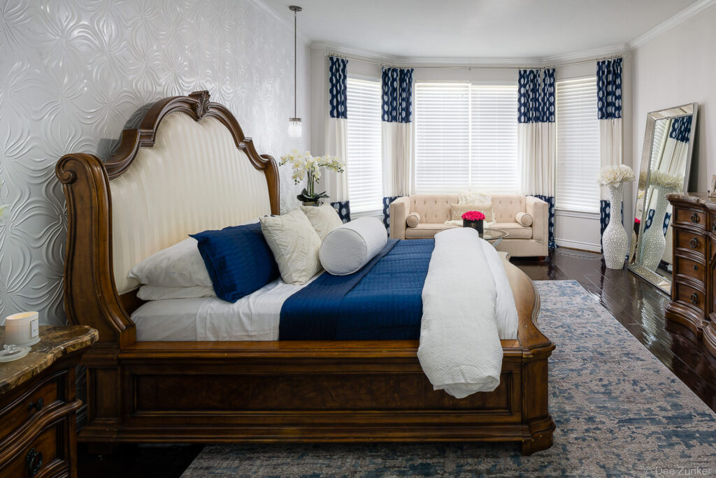 Ornate king bed with navy and white bedding in a luxury Houston master bedroom featuring bay windows and tufted seating area.