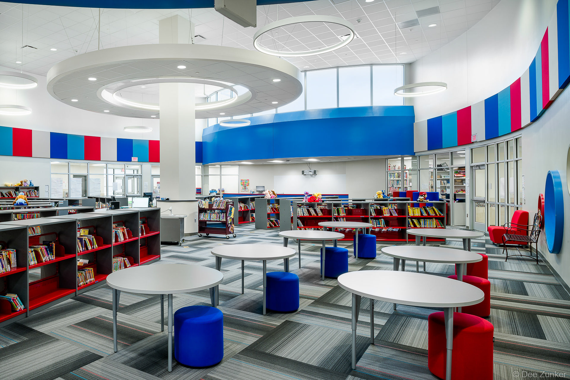 Bright elementary school library interior with round tables, blue and red seating, book shelves, and modern circular ceiling lights