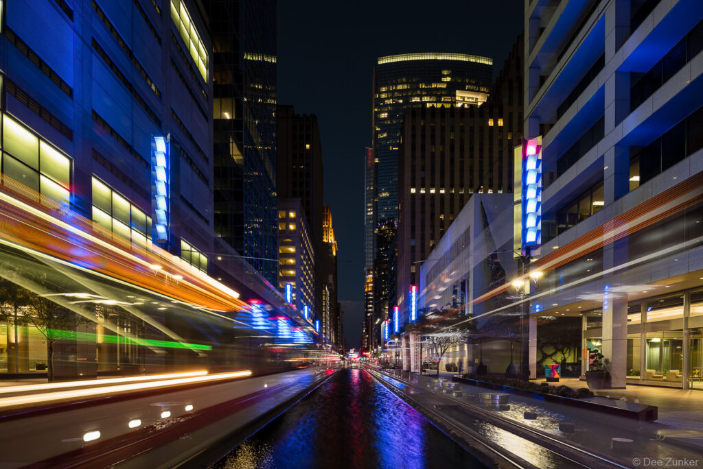 Long exposure night photograph of Main Street Square Houston with light trails from passing trains and glowing blue facade lighting