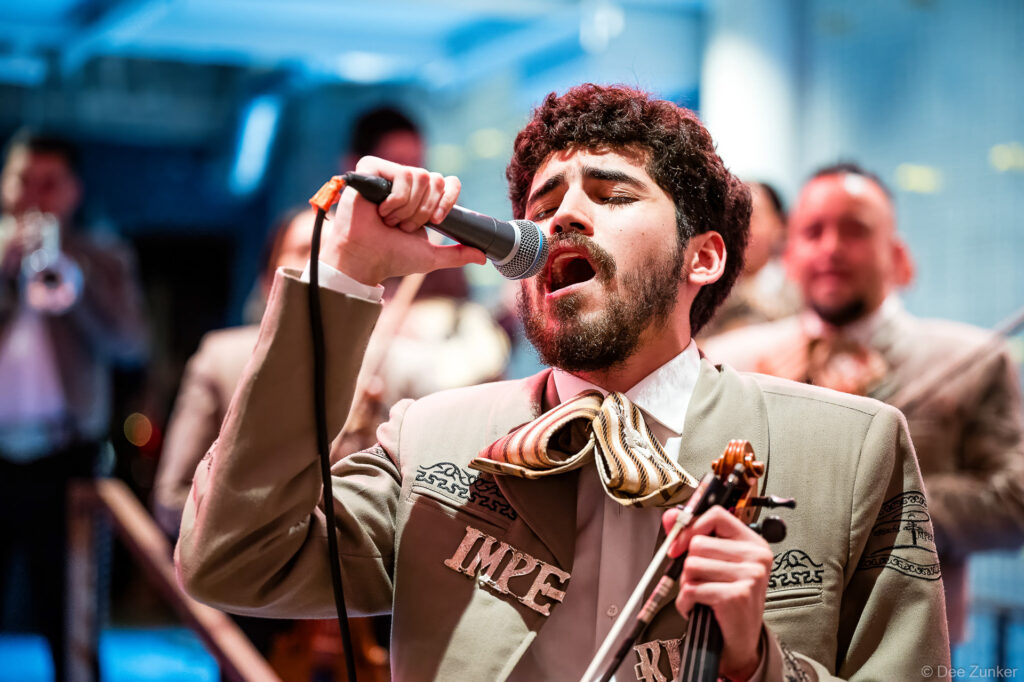 Passionate mariachi vocalist singing into microphone holding violin at Houston corporate event, wearing traditional charro suit.