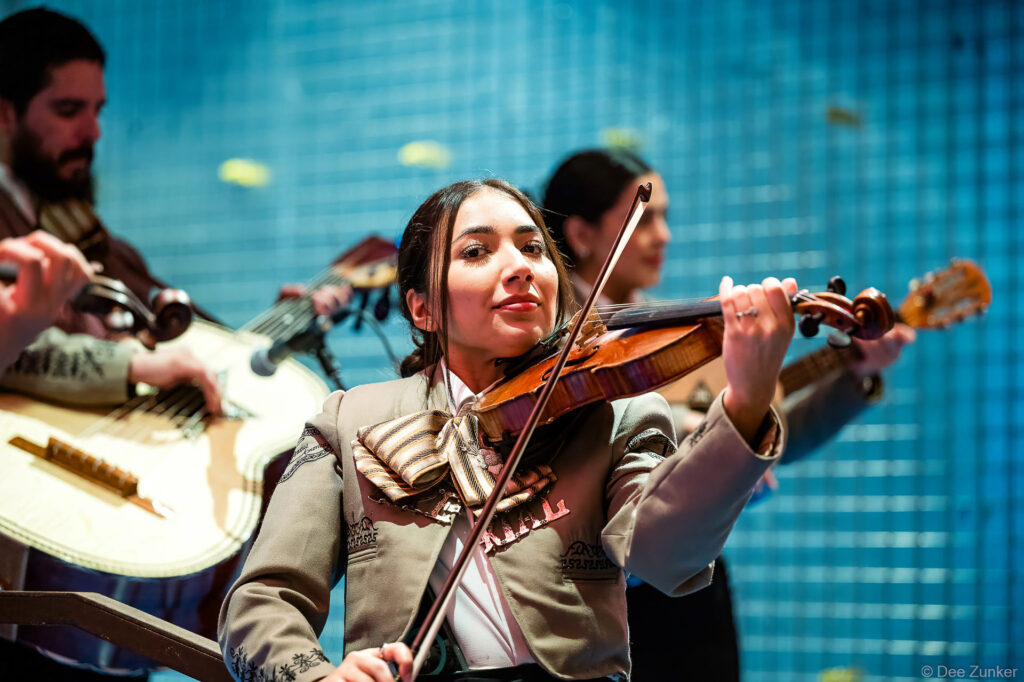 Female mariachi violinist in charro suit performs at Houston corporate gala, with guitarist visible in background.