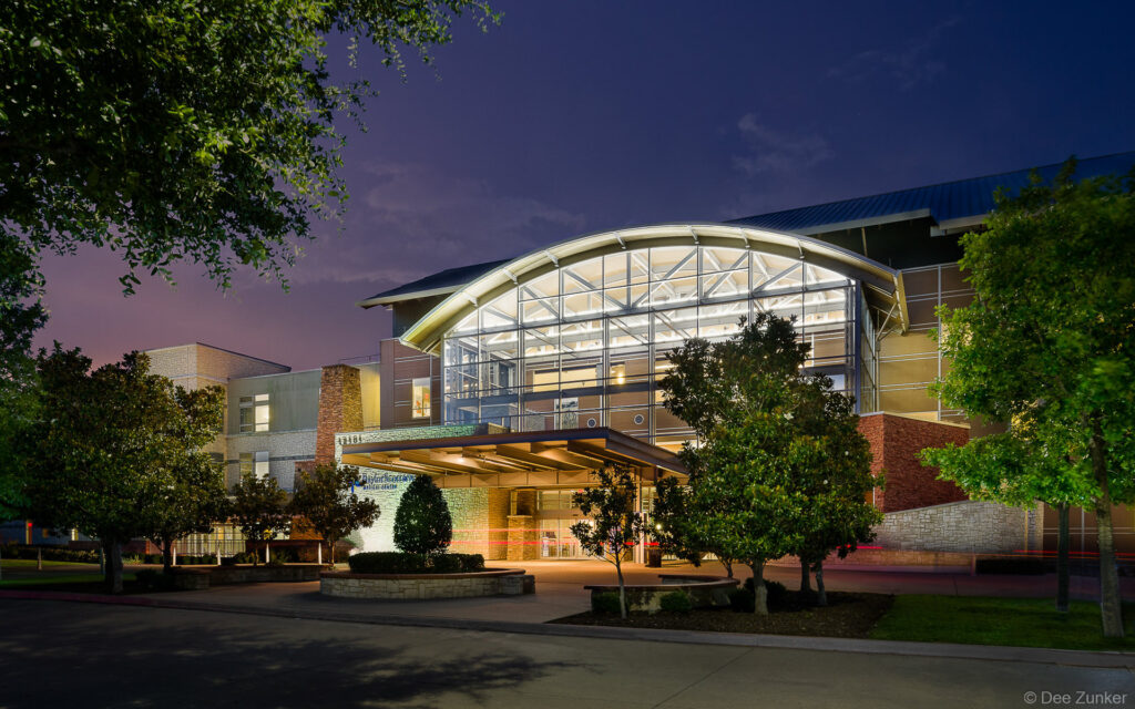 Medical center exterior at twilight featuring illuminated glass facade arched entrance landscaped grounds Plano Texas architectural photography