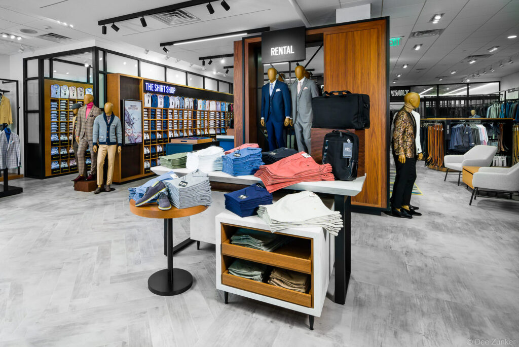 Modern menswear retail store interior featuring rental suits, shirt shop, folded casual pants display, and mannequins in formal attire, Houston TX.