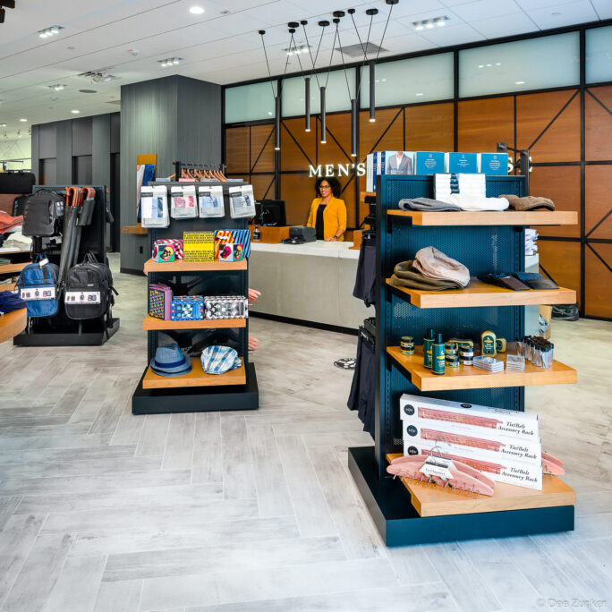 Modern men's retail department interior with accessory display shelves, hats, boxed merchandise, and a sales associate at the checkout counter.