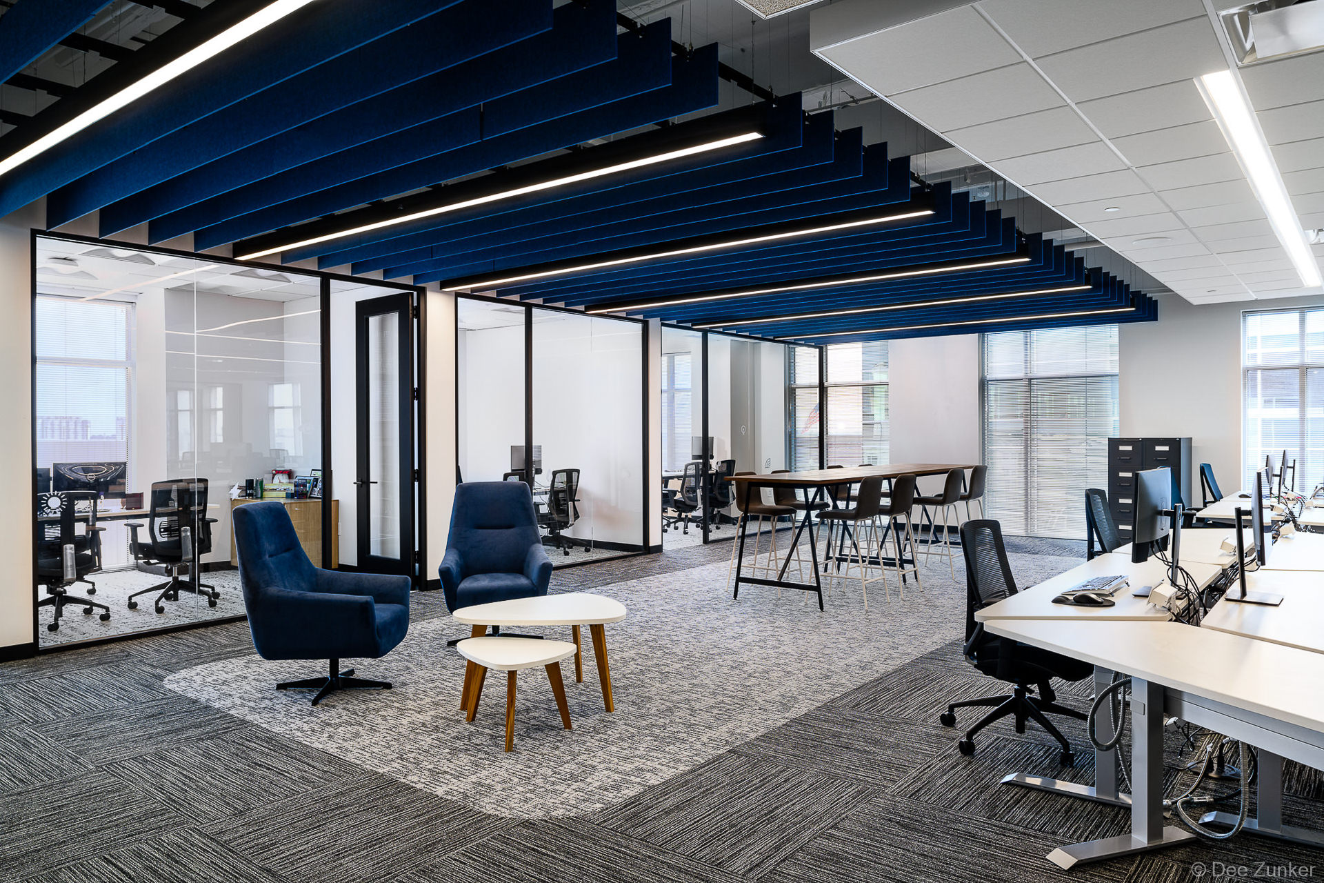 Modern open-plan corporate office featuring blue lounge chairs, collaborative seating areas, glass partitions, and blue acoustic ceiling panels in Houston