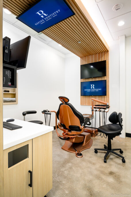 Copper dental chair in a modern Houston dentistry treatment room with wood slat accent wall, dual monitors, and dental equipment.