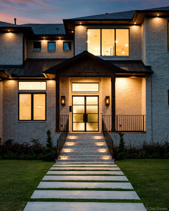 Illuminated modern brick home exterior at dusk with lit staircase, black-framed glass entry door, and manicured lawn in Houston TX.