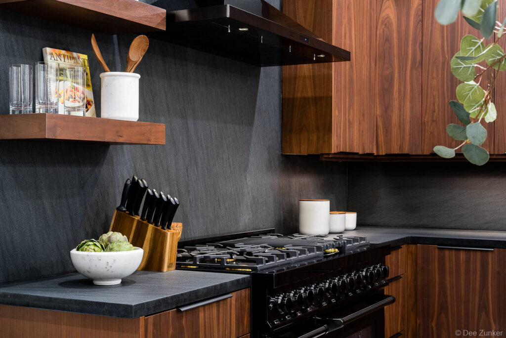 Luxury kitchen interior with dark slate backsplash, walnut cabinets, black gas range, knife block, and open shelving styled with cookbooks and glassware.