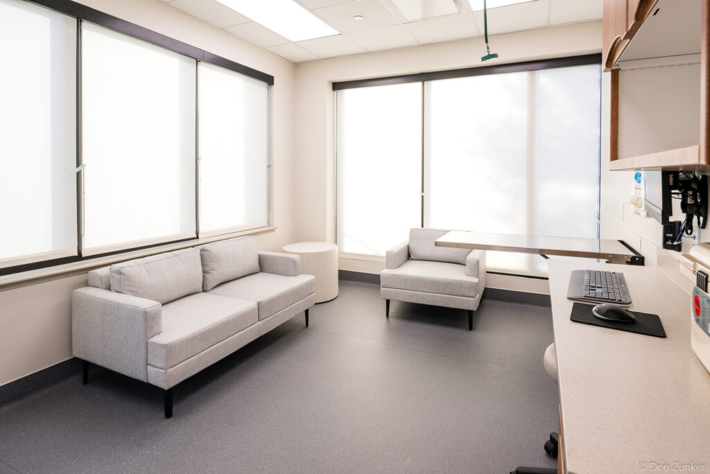 Bright veterinary consultation room with gray sofas, armchair, workstation, and roller-shade windows in a modern Houston clinic interior.
