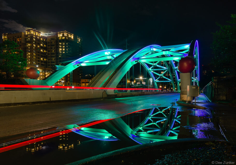 Montrose Bridge glowing teal and blue at night reflected in rain puddles with Houston skyline in background
