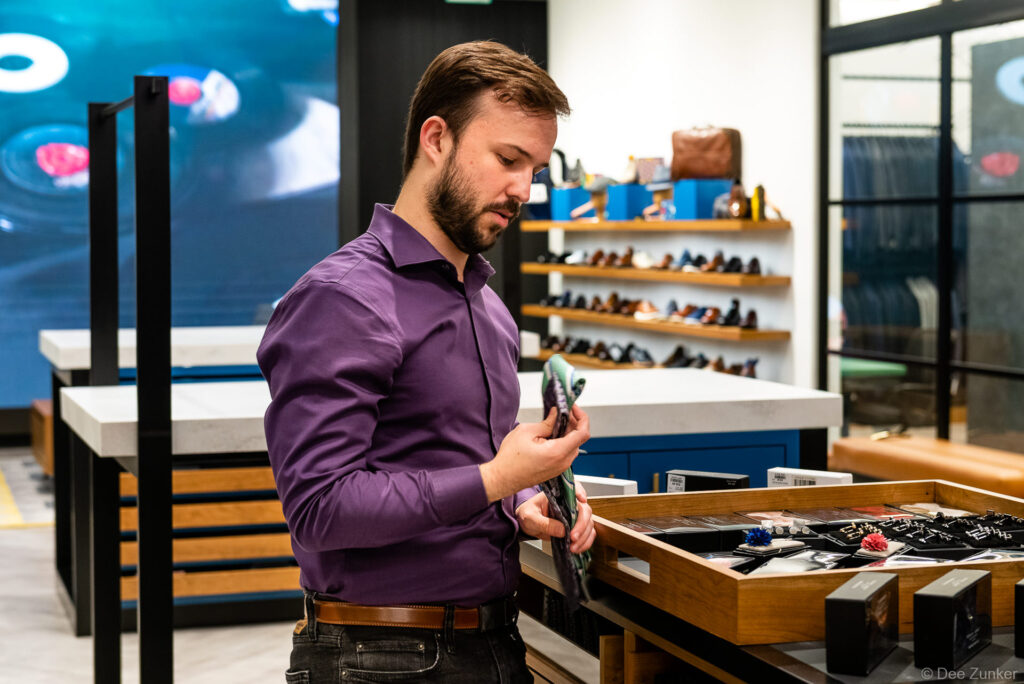 Bearded man in purple shirt examines accessories at a wooden display table inside a modern retail clothing and shoe store.