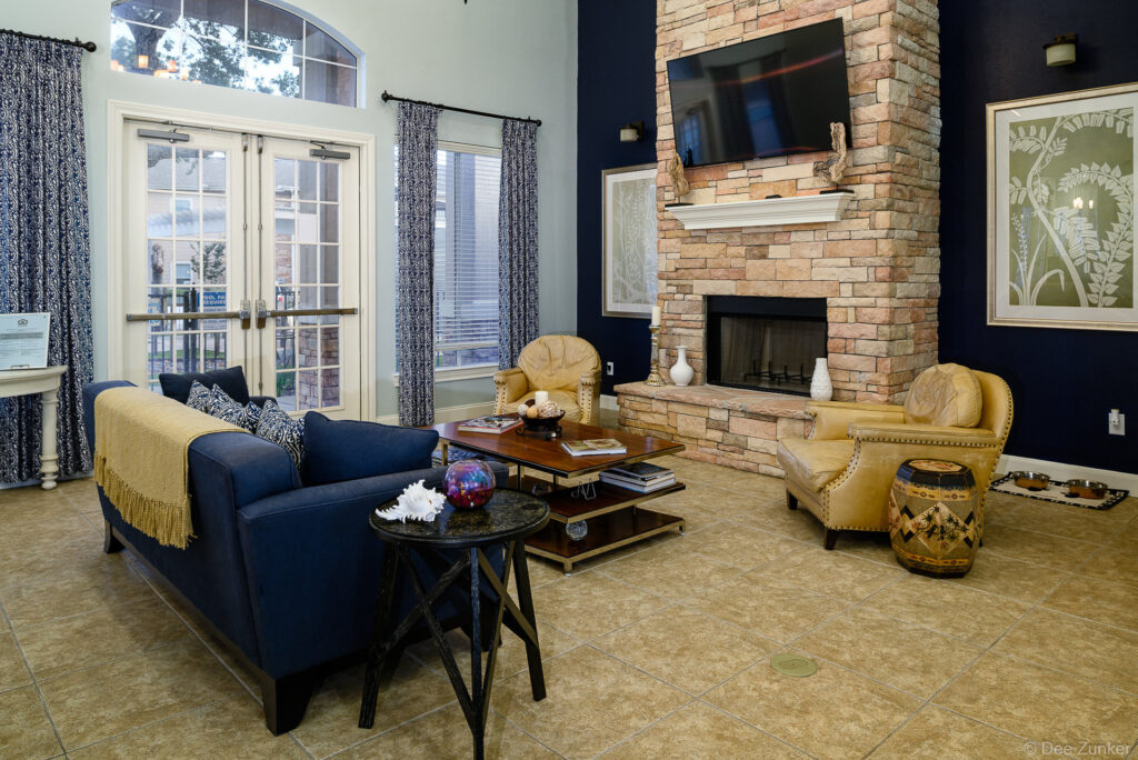 Stylish apartment clubhouse lounge with navy sofa, stone fireplace, flat-screen TV, and gold accent chairs at Oaks Bentwater Houston.