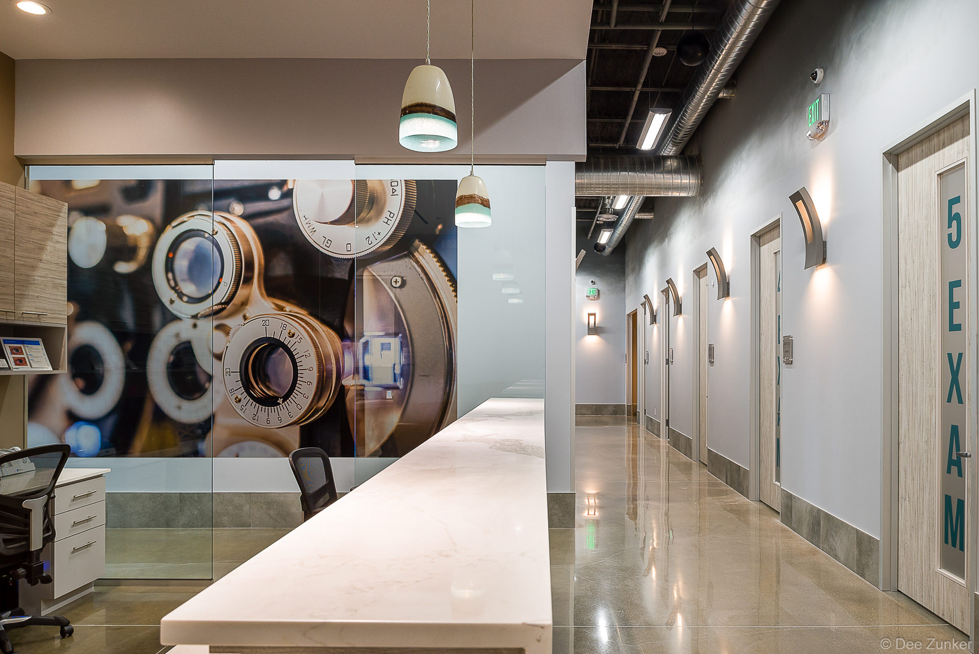 Modern optometry clinic hallway with exam rooms, marble reception counter, phoropter wall mural, and industrial-style lighting in Houston.