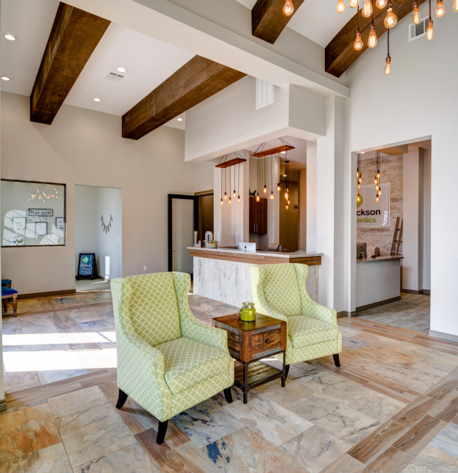 Bright orthodontic office waiting area with green wingback chairs, wood beam ceiling, Edison bulb pendants, and stone tile flooring