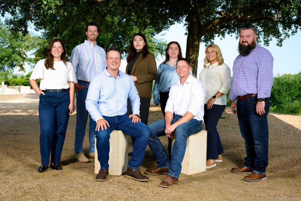 Nine corporate team members posed outdoors under oak trees on a gravel path for a Houston company group portrait session.