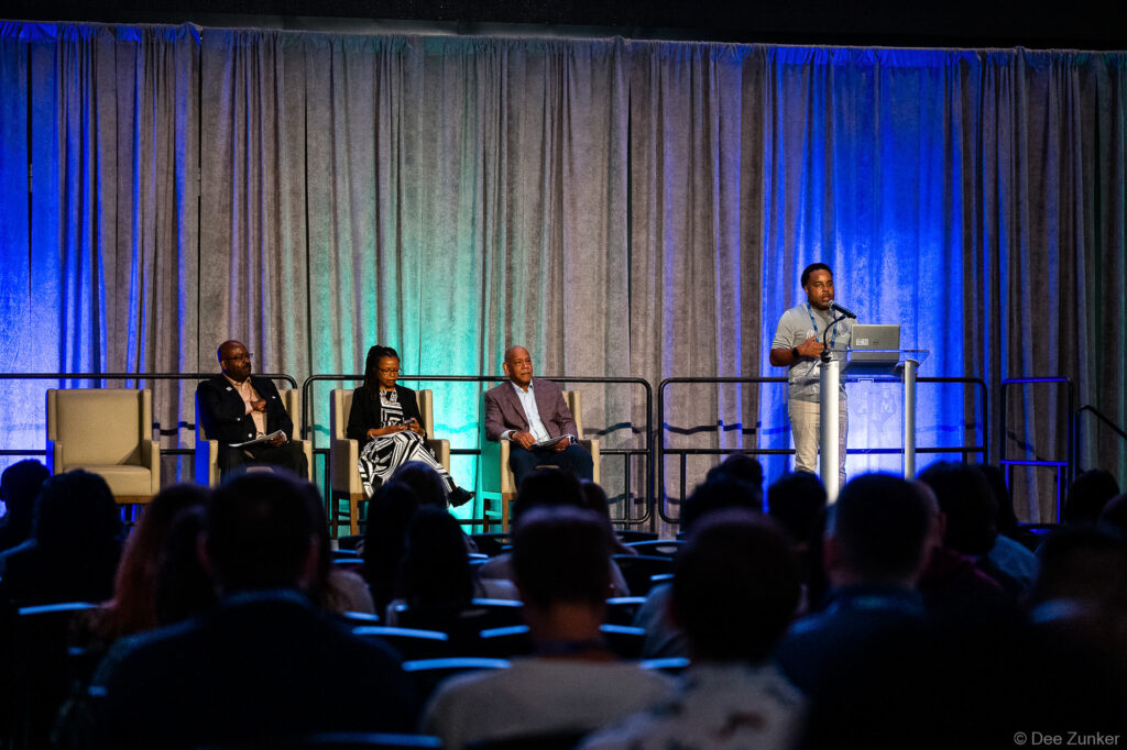 Four panelists on a blue-lit conference stage, three seated and one speaking at a podium before a packed audience in Houston.