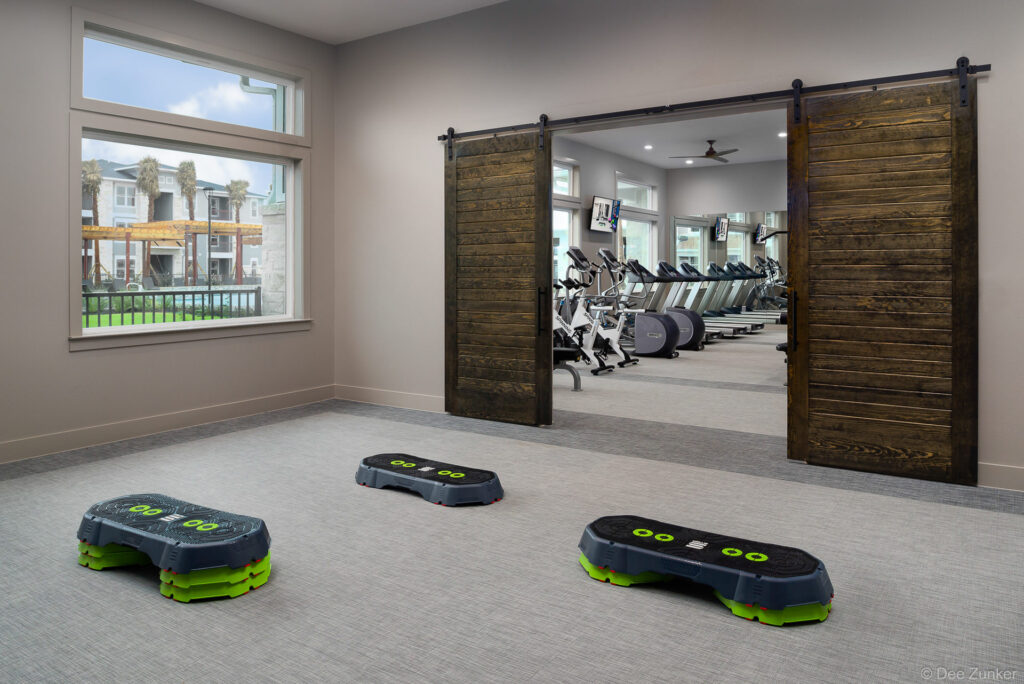 Apartment fitness center aerobic studio with step platforms and adjacent cardio room featuring treadmills, visible through rustic barn doors