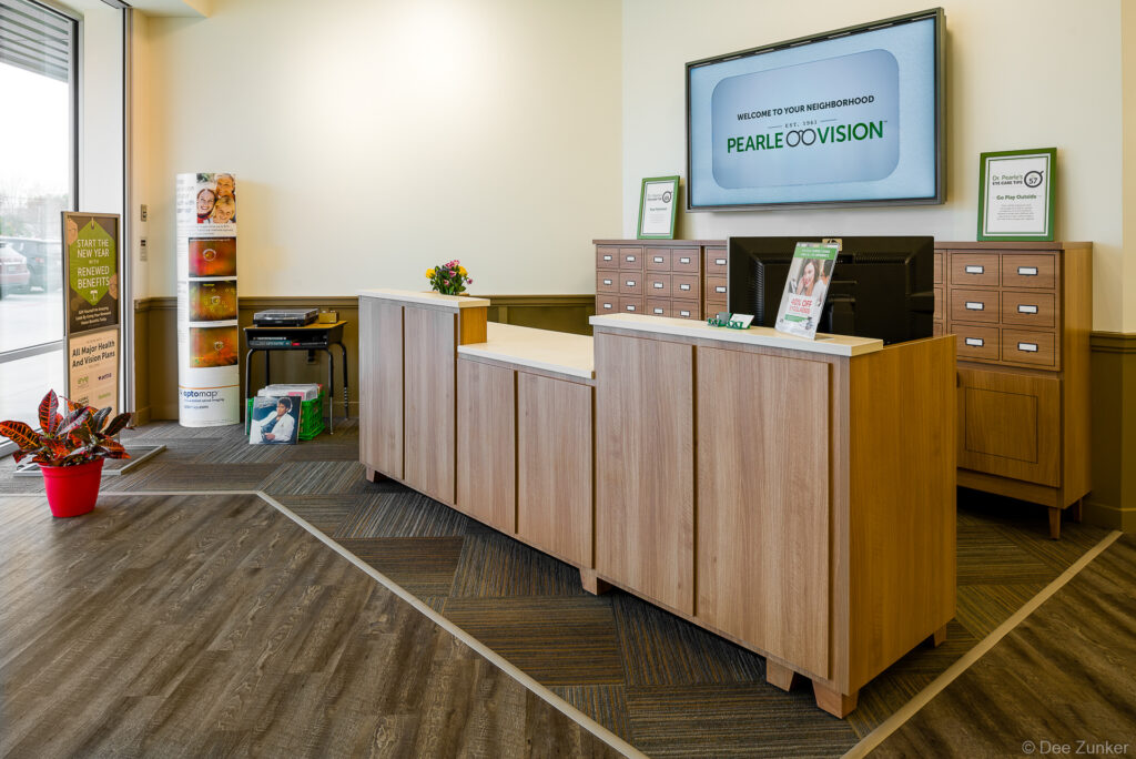 Modern wood reception desk at a Pearle Vision optometry office with welcome screen, framed displays, and floral arrangement in Houston, TX.