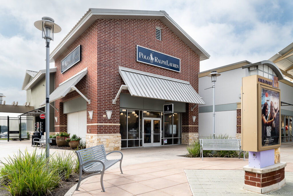 Exterior of a brick polo ralph lauren outlet store at a Cypress TX shopping center, featuring metal awnings, landscaped plaza, and directory signage.