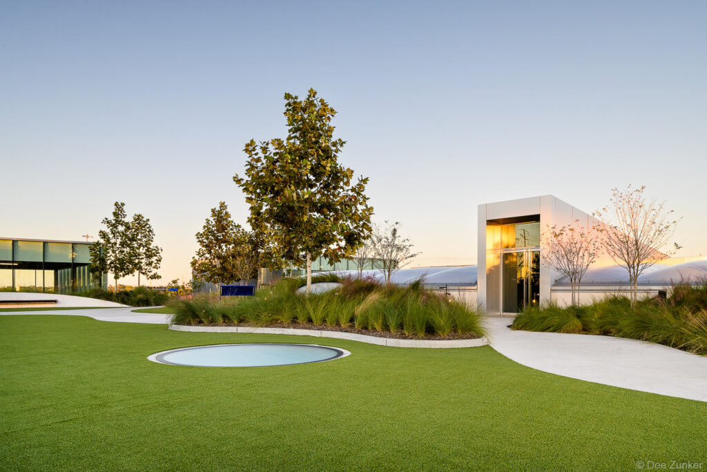 Rooftop garden at POST Houston featuring lush green lawn, circular skylight, ornamental grasses, and modern architectural pavilion at dusk