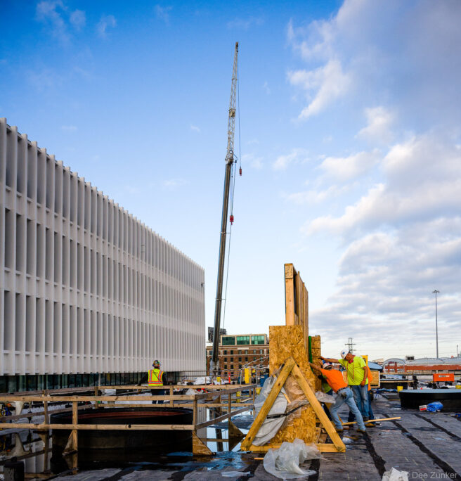 Construction workers removing wood forms from glass on a rooftop deck at Post HTX Houston with a crane and adjacent parking structure visible.