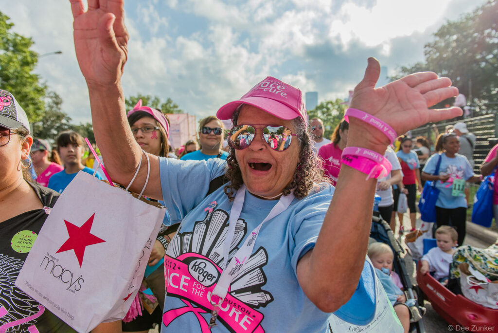 Excited woman in pink cap and sunglasses cheers with arms raised at Houston Race for the Cure corporate event.