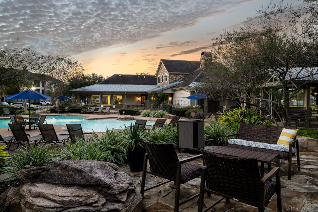 Resort-style apartment pool area at twilight with wicker lounge seating, lush landscaping, and illuminated clubhouse in Pearland Texas