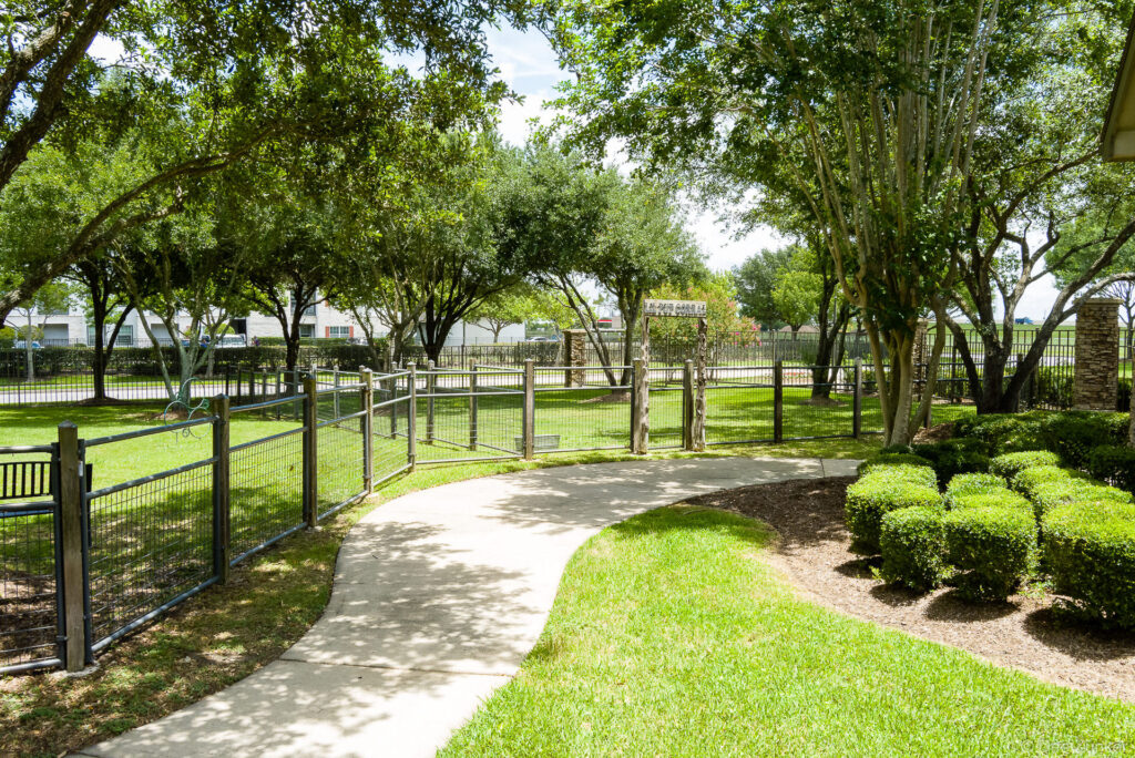 Curved concrete pathway leads through a fenced dog park with mature trees and manicured landscaping at Ranch City Park, Houston.