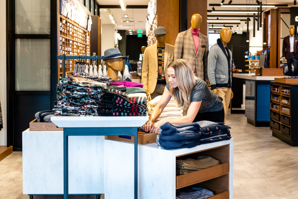 Female retail associate arranges folded menswear on a display table inside a well-styled Houston menswear boutique.