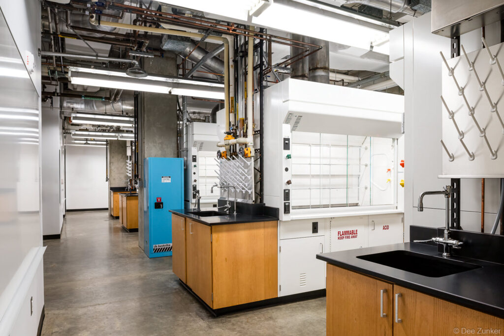 Renovated university chemistry lab interior featuring fume hoods, glassware drying stations, wood cabinetry, and exposed industrial ceiling systems