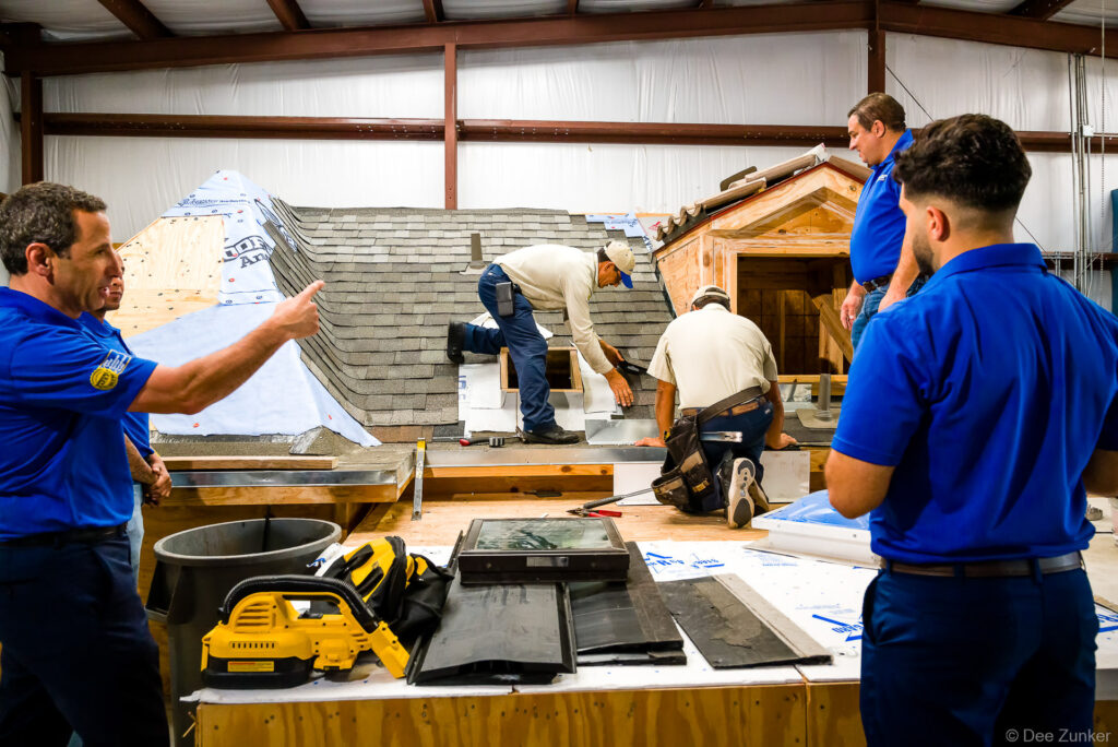 Roofing contractors install shingles on an indoor training mock-up roof while supervisors in blue polo shirts observe, Houston TX.