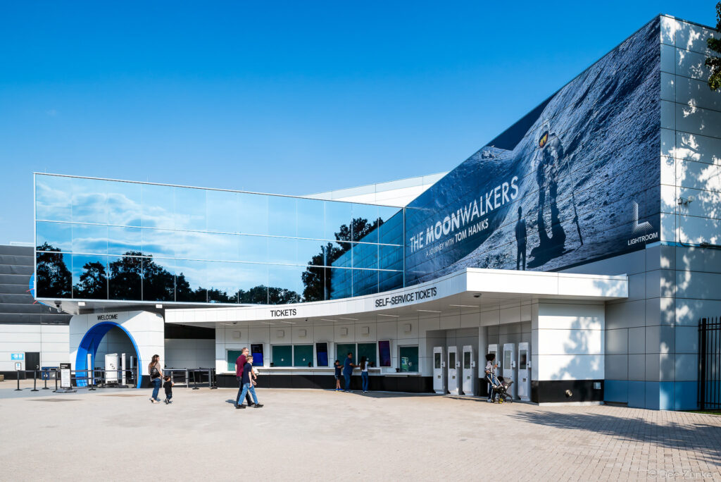 Exterior entrance and self-service ticket counter at Space Center Houston featuring large Moonwalkers exhibition signage on a clear sunny day