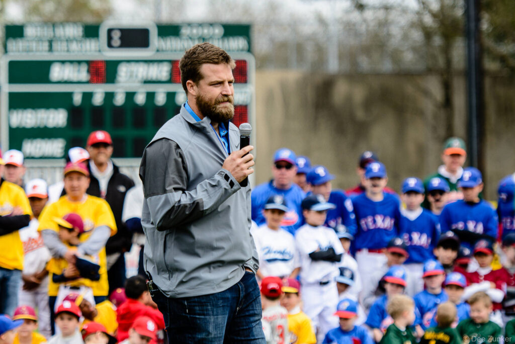 Bearded male speaker holds microphone at youth baseball league opening day ceremony with players in Texas Rangers uniforms behind him.