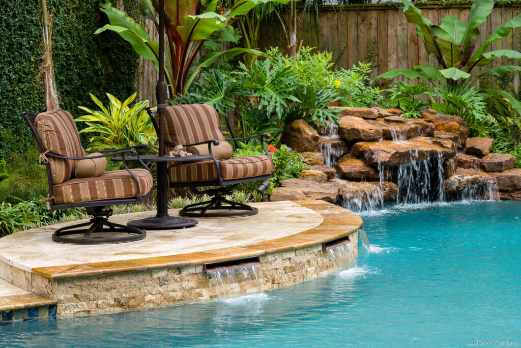 Luxury backyard pool at Sugar Hill Houston featuring a rock waterfall, tropical landscaping, and wrought iron patio chairs on a stone deck.